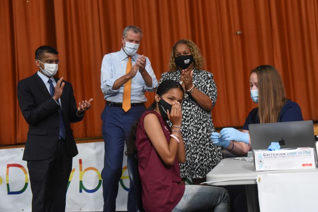 A girl clasps her hands in front of her face after getting a vaccine in a school auditorium with the Mayor, Health Commissioner, and Schools Chancellor looking on, as well as as nurse